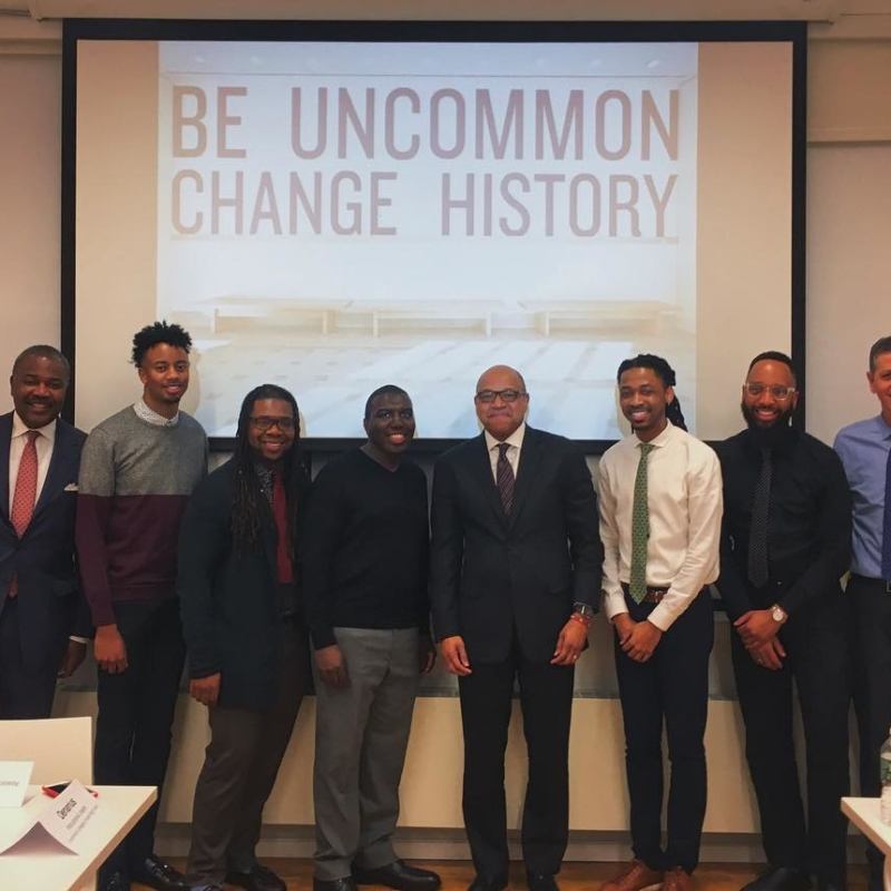 Group of men stand in front of projection that reads "Be Uncommon Change History"