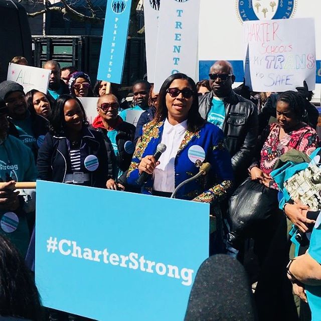 Woman speaks into microphone behind sign that reads "#CharterStrong"
