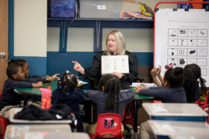 Teacher sitting in chair with students surrounding her as she reads to them.