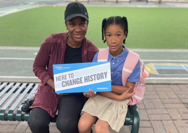 Parent and child smiling and holding a "Here to Change History" sign on the first day of school